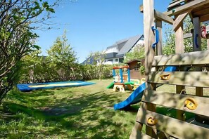 Eine hölzerne Spielkonstruktion mit Rutschen und Kletterelementen, eine blaue Rutsche und ein kleines Spielhaus in einem Garten.