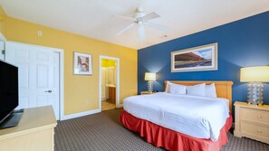 Ein Hotelzimmer mit Bett, Fernseher, Schreibtisch, Deckenventilator und einem Bild an der Wand.
