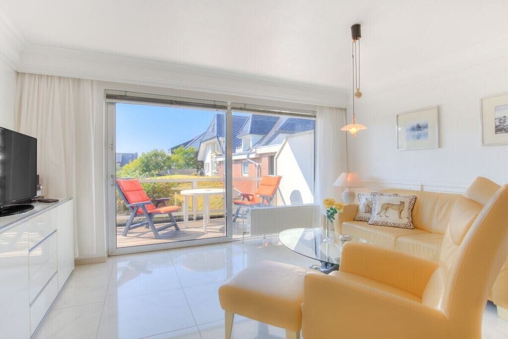 Un salon moderne avec un canapé, une table basse en verre et un téléviseur. Une porte-fenêtre coulissante donne accès à un espace extérieur aménagé avec une table et des chaises.