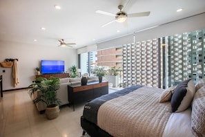 A modern bedroom with a large bed, a TV, a desk, and a balcony with a view of a high-rise building. - Infinity Pool & Hot Tub | Romantic Zone Hideaway (Puerto Vallarta)
