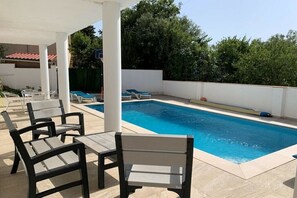 Ein Schwimmbecken mit Liegestühlen und einem Tisch, umgeben von einem weißen Zaun und viel Grün.