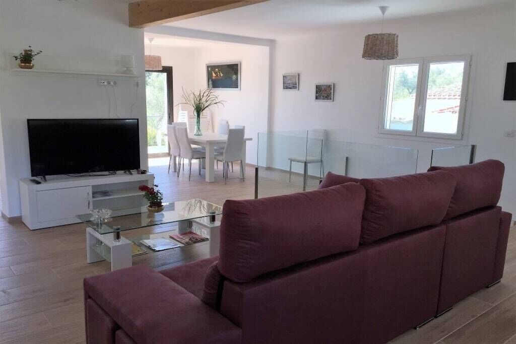 A modern living room with a flat-screen TV, a glass coffee table, and a red sofa. There is a dining area with a white table and chairs visible through an open doorway.
