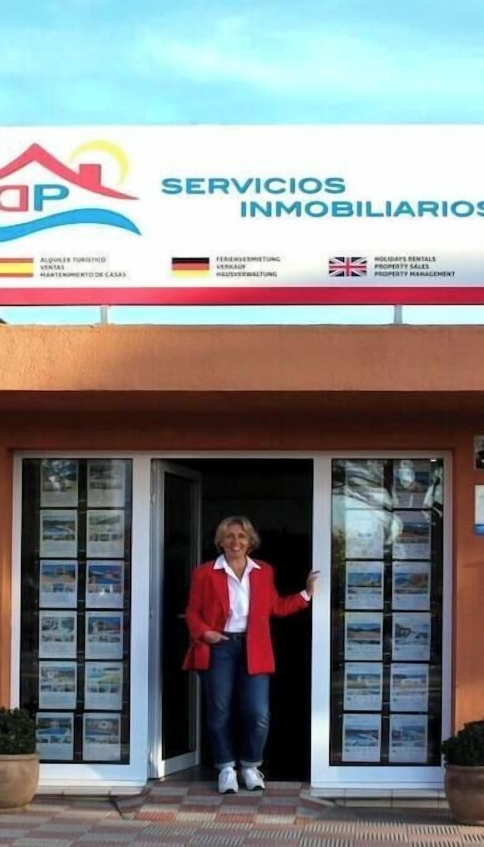 Una persona de pie frente a la entrada de un edificio con un cartel que dice 'SERVICIOS INMOBILIARIOS' y banderas de España, Alemania y el Reino Unido.