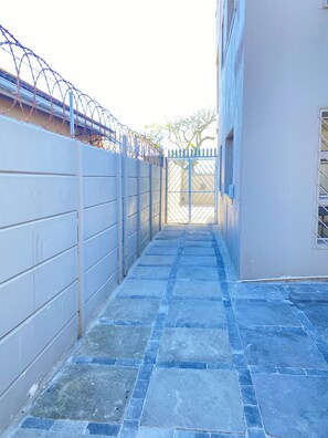 Un sendero estrecho que conduce a una entrada con portón, una cerca azul y un edificio a la derecha.