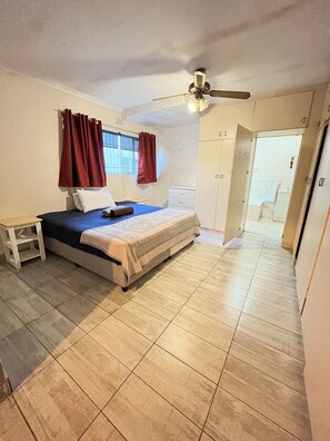Un dormitorio con cama, mesita de noche, ventilador de techo y ventana con cortinas.