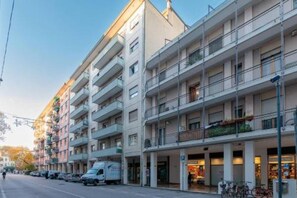 Um edifício residencial com vários andares, varandas, um espaço comercial no térreo e carros estacionados na rua.