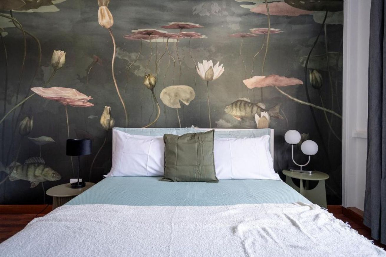 Un dormitorio con cama, almohadas, una mesita de noche, una lámpara y un mural en la pared.