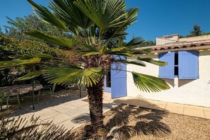 Um edifício com persianas azuis, uma palmeira e um alpendre com cadeiras e uma mesa.