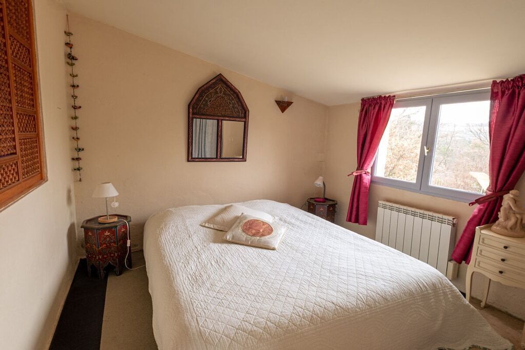 Ein Schlafzimmer mit einem großen Bett, einem Fenster mit Vorhängen, einem Heizkörper und einem an der Wand befestigten Spiegel.