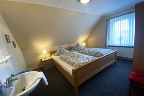 Een slaapkamer met een bed, nachtkastje, lamp, raam met gordijnen en een badkamer met wastafel.