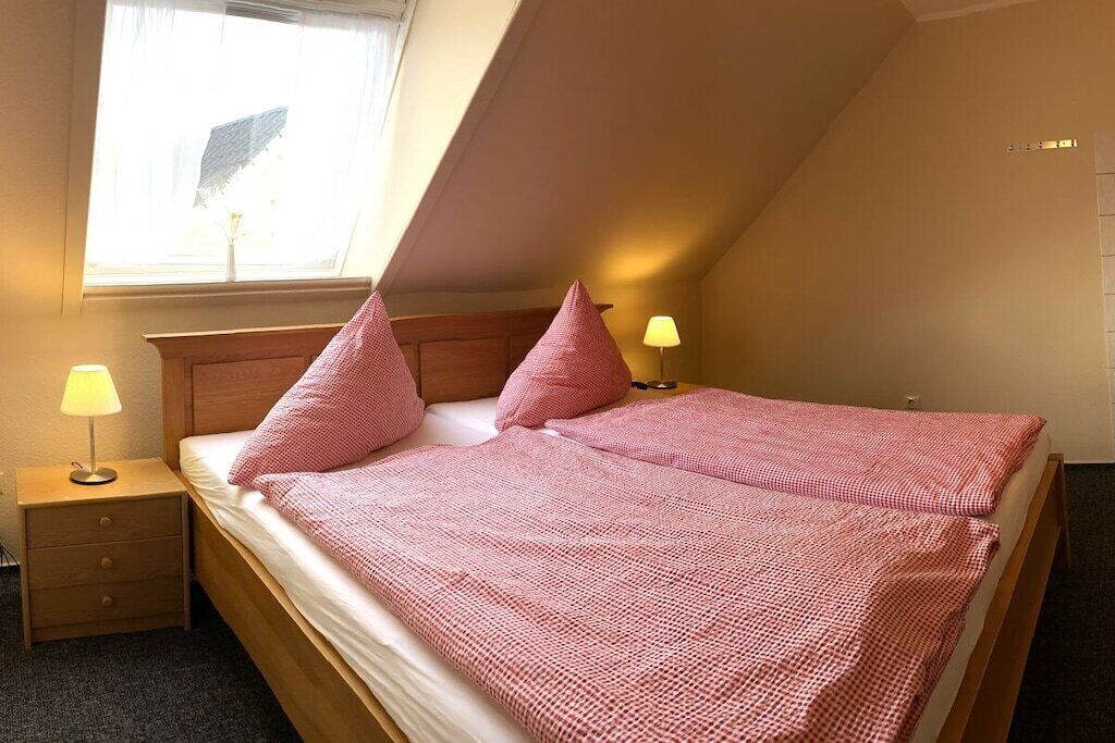 Une chambre avec un plafond en pente, un lit recouvert d’une couverture à carreaux roses, deux oreillers, une table de chevet avec une lampe et une fenêtre laissant entrer la lumière naturelle.