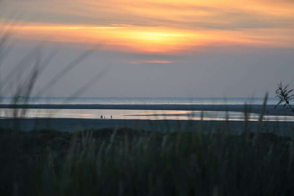 Escena de playa con atardecer, mar tranquilo y figuras a lo lejos.