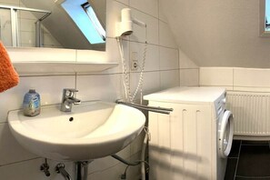 Een badkamer met een wastafel, een spiegel, een handdoek, een fles handzeep, een douchekop en een wasmachine.