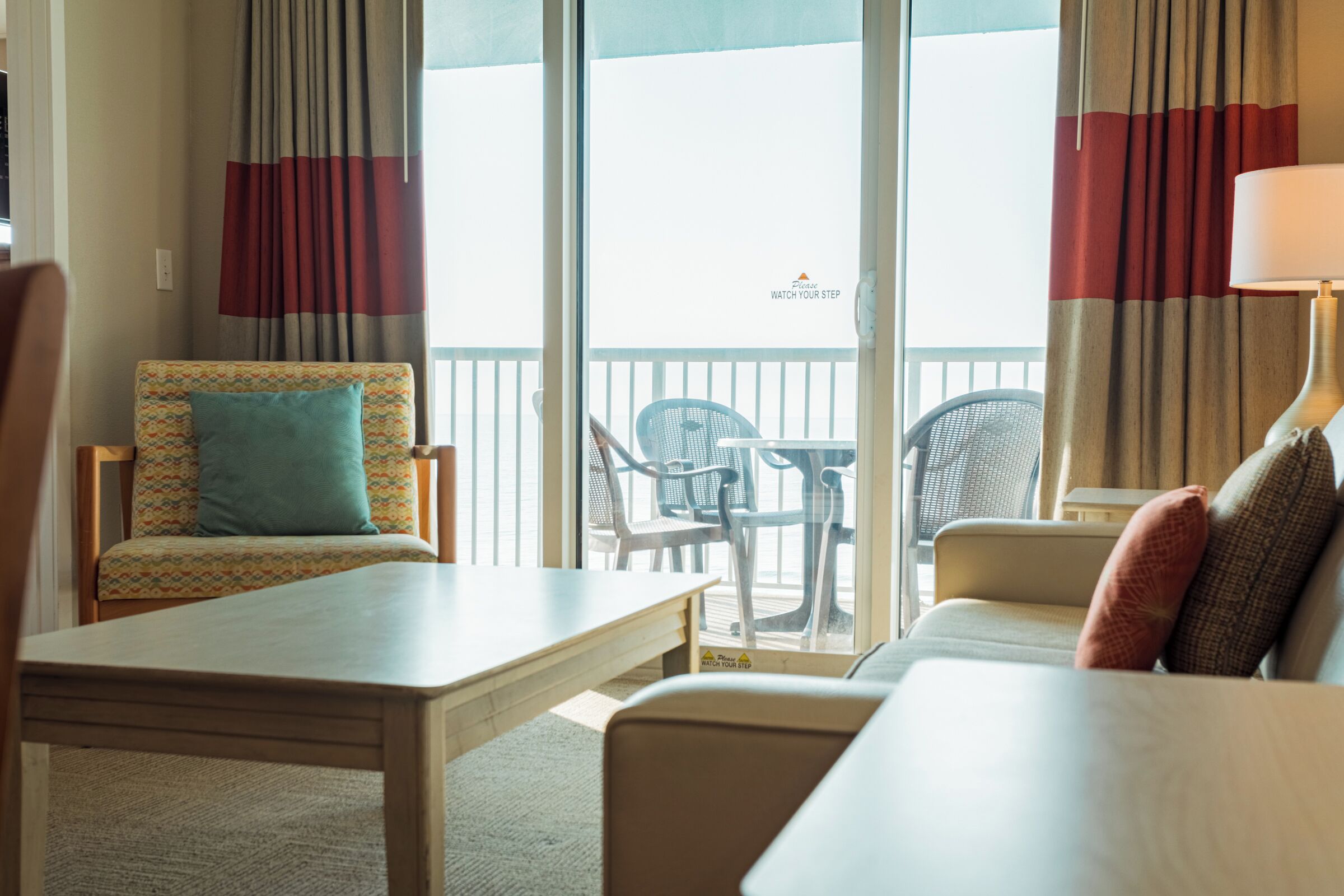 A hotel room with a balcony, a sofa, a chair, a table, and a lamp.
