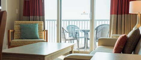 A hotel room with a balcony, a sofa, a chair, a table, and a lamp.