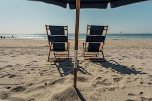 Une plage avec deux chaises de plage en bois sous un grand parasol sombre, une plage de sable et l’océan en arrière-plan.