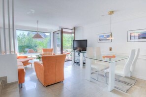 Een moderne woonkamer met een glazen eettafel, oranje banken en een flatscreen televisie.