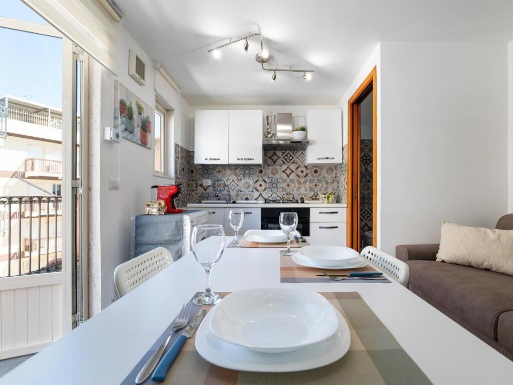 A dining area with a table set for two, a kitchen in the background, and a balcony with a railing.