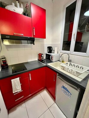 A compact kitchen with red cabinets, a black countertop, a built-in stove, a microwave, a coffee maker, and a sink with a white basin. - Sweet Home Montmartre / 2® / Eugène Süe* (Paris)