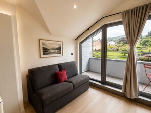 Un salón moderno con un sofá gris oscuro, un cojín rojo, una fotografía enmarcada de un paisaje en la pared y un amplio ventanal corredera que da a un balcón con vistas a un paisaje verde.