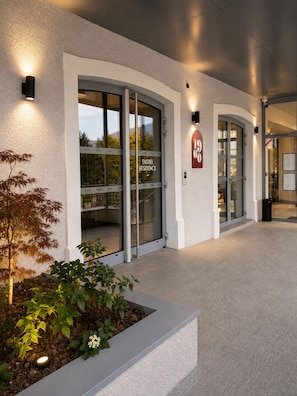 A hotel entrance with glass doors, a sign indicating room number 1900, and a landscaped area with plants.
