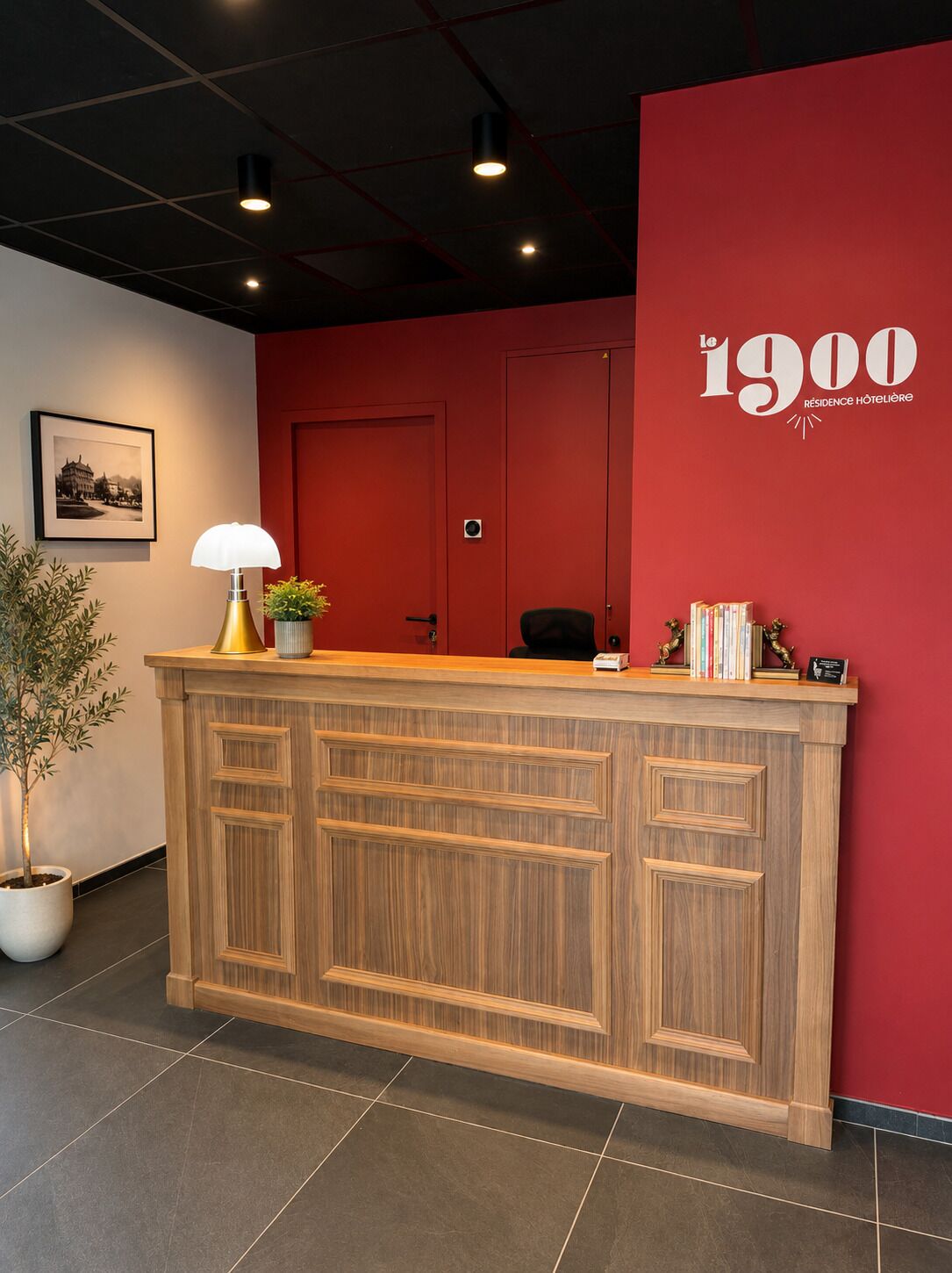 A reception desk with a red wall, a framed picture, a potted plant, and a lamp.