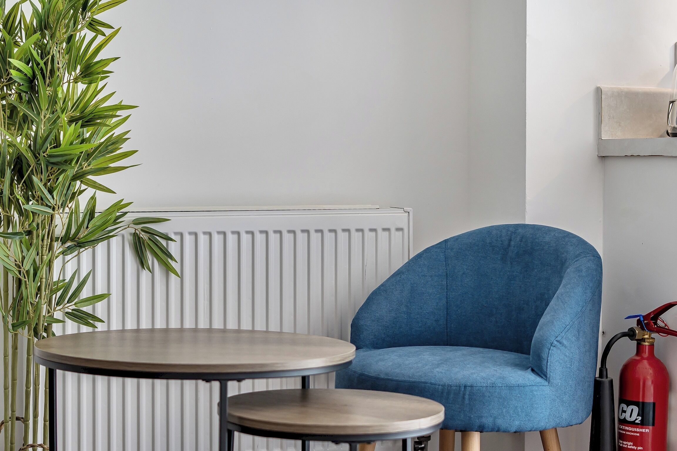 A room with a blue armchair, a small round table, a radiator, and a fire extinguisher on the wall.