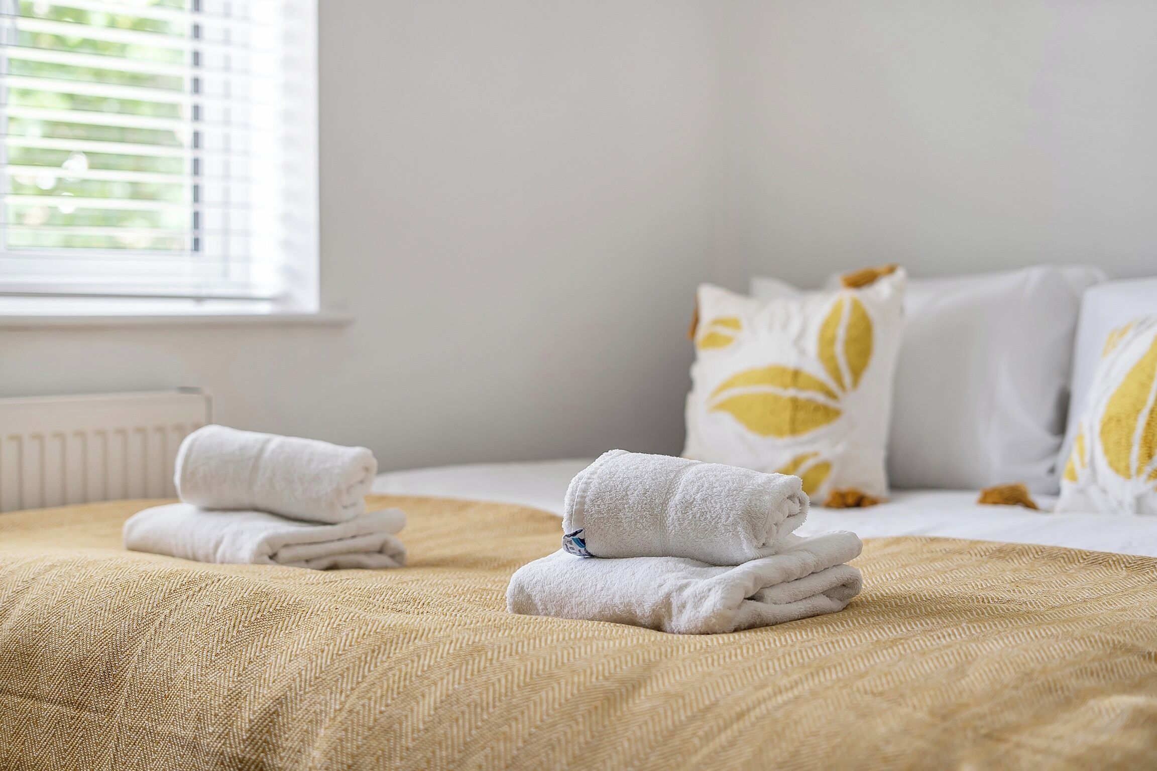 A neatly made bed with folded towels, a window with blinds, and a bed with white and yellow pillows.