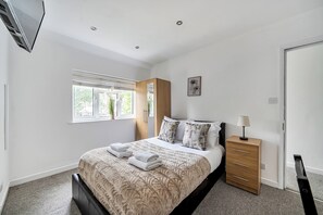 A bedroom with a bed, a bedside table, a wardrobe, a window with blinds, and a wall-mounted air conditioning unit. - Surrey 5-Bed Home for Groups & Business Stays - Pass the Keys (Addlestone)