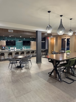 Un comedor moderno con una mesa grande y sillas, una cocina al fondo, e iluminación colgante.