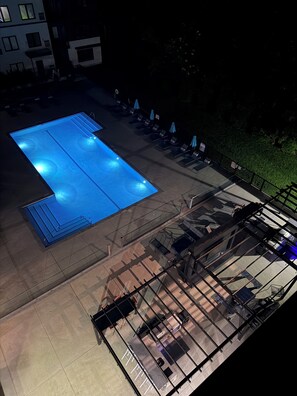 A rooftop pool area with a blue-lit pool, lounge chairs, and a glass railing.