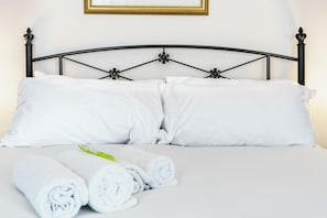 Cama bem arrumada com lençóis e almofadas brancas, toalha branca enrolada e um quadro na parede.