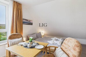 A hotel room with a bed, a dining table with plates and cups, a window with curtains, and a wall-mounted sign with 'LIG'. - Holiday apartment Gorch Fock 18 (Langeoog)