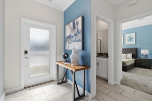 A room with a door, a console table with a globe and a decorative item, a blue accent wall, a bed with a nightstand, and a bathroom with a mirror and sink. - Spacious Champions Gate Townhome - Splash Pool - Walk to Resort - Near Disney (Kissimmee)