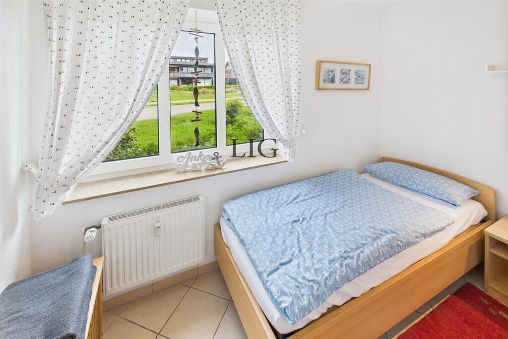 A bedroom with a bed, a radiator, a window with curtains, and a small shelf.