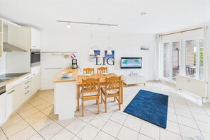 Eine moderne Küche mit weißen Schränken, einem Essbereich mit Holzsesseln, einem blauen Teppich und einem Fernseher an der Wand.