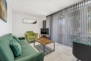 Een moderne woonkamer met een groene bank, een groene fauteuil, een salontafel, een televisie en een groot raam met zonwering.