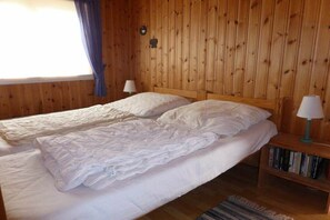 Ein Zimmer mit zwei Betten, Holzwänden, einem Fenster mit Vorhängen, einem Nachttisch mit Lampe und einem Bücherregal.
