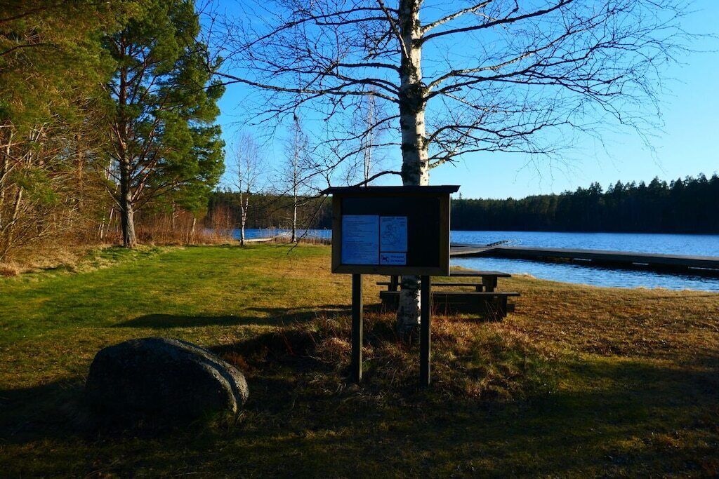 Een houten informatiebord bij een meer, met een picknicktafel en een grote rots op de voorgrond.