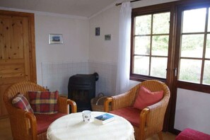 Ein gemütliches Zimmer mit Korbstühlen, einem kleinen Tisch mit Buch und Tasse, einem Kamin und einem Fenster mit Aussicht.