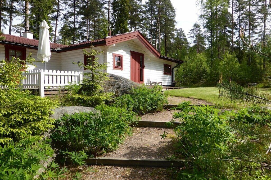 Une maison de plain-pied avec une porte rouge et des murs blancs, entourée d’une clôture en bois peinte en blanc, située dans une zone boisée et accessible par un chemin de gravier menant à l’entrée.
