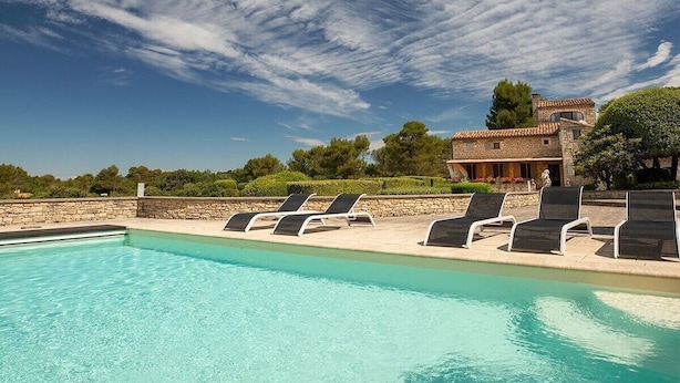 Une piscine avec des chaises longues, un mur en pierre et, au fond, un bâtiment avec un toit recouvert de tuiles.