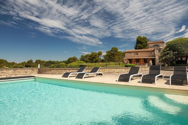 A swimming pool with lounge chairs, a stone wall, and a building with a tiled roof in the background.