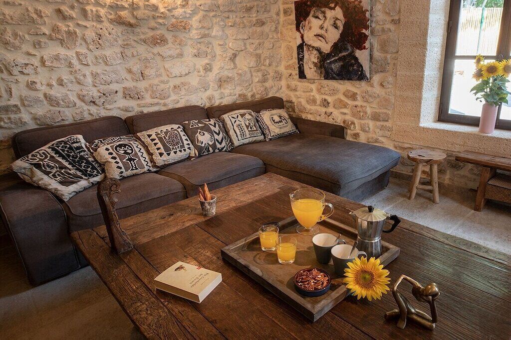 Un salon chaleureux avec un mur en pierre, un canapé gris agrémenté de coussins à motifs, une table basse en bois sur laquelle sont disposés des éléments pour le petit-déjeuner, et un tableau accroché au mur.