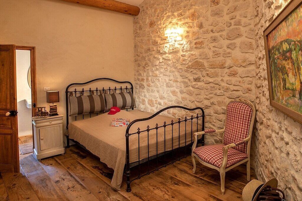 A bedroom with a stone wall, a bed with a metal frame, a chair, and a small bedside table with a lamp.