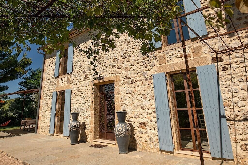 Ein Steinhaus mit rustikalem Aussehen, ausgestattet mit großen Fenstern und Fensterläden, einer Tür mit einem Metallgitter und zwei großen Vasen im Vordergrund.