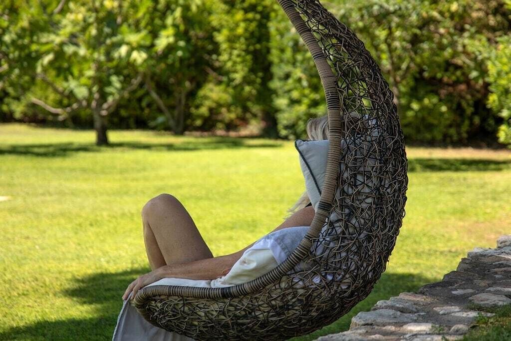 A person reclining in a wicker chair outdoors with a grassy area and trees in the background.