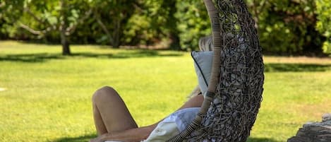 Una persona reclinada en una silla de mimbre al aire libre, con un césped y árboles de fondo.