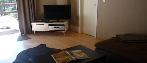 A room with a television on a stand, a small table with a magazine, a sofa, and a door.