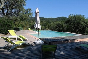 Ein Swimmingpool mit Liegestühlen und einem Sonnenschirm auf einer Holzterrasse, umgeben von Bäumen und Hügeln.